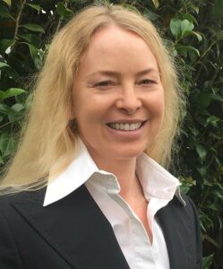 A woman in a black suit smiling in front of bushes.