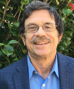 A man with glasses and a mustache in front of bushes.