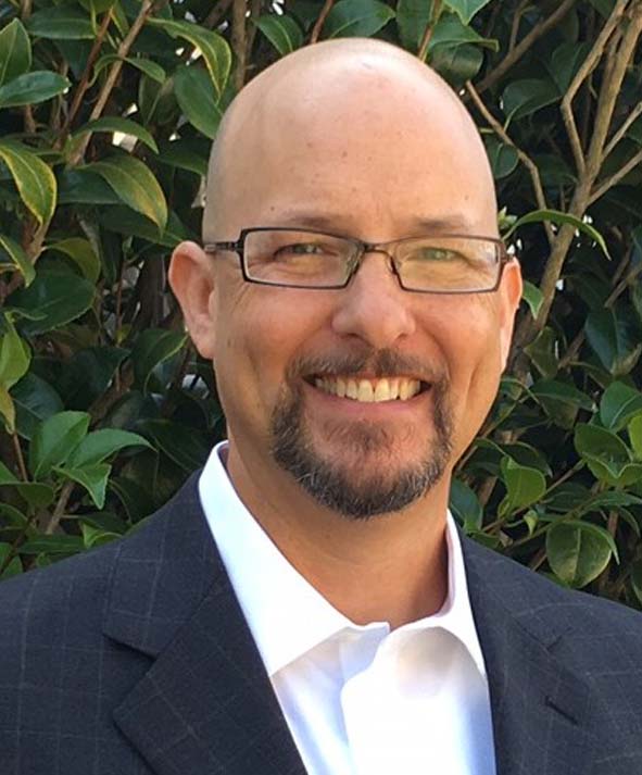 A bald man smiling in front of bushes.