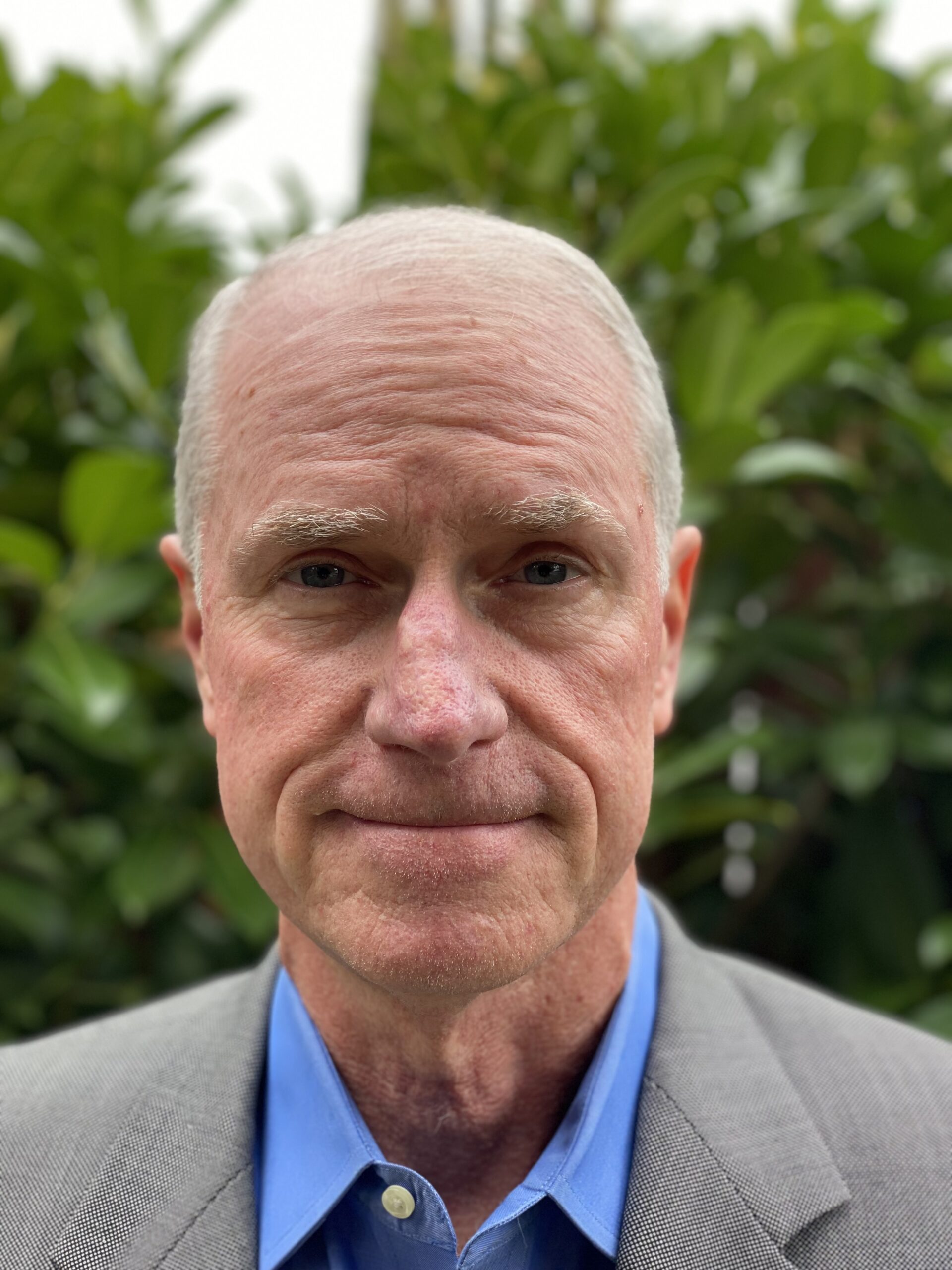 An Additional Advisor, dressed in a suit, is standing confidently in front of some lush green bushes.