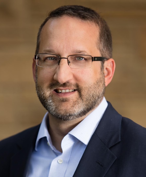 A confident man with glasses and a beard displaying successful management skills, smiling in front of a brick wall.